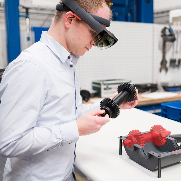 Mitarbeiter mit AR-Brille prüft ein schwarzes Bauteil an einem Arbeitstisch in einer Werkstatt