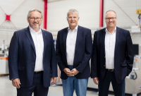 Drei Männer in dunklen Anzügen stehen nebeneinander vor einer hellen Hallenkulisse mit roten Elementen