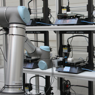 Kollaborativer Roboterarm vor einem Regal mit mehreren 3D-Druckern in einer automatisierten Laborumgebung